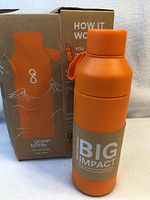 Front view of orange metal water bottle with cardboard sleeve, displayed alongside its box which shows usage instructions and branding.