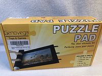 Box front showing Genovega Puzzle Pad kit with text and illustration of puzzle mat with hot air balloons image, rolled with storage tube.