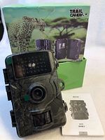 Trail camera resting against its packaging box and instruction manual, showing the front of the camera with night vision LEDs and lens, camouflage pattern, and box featuring a cheetah image.