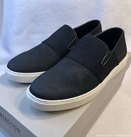 Side angle view of black slip-on loafers with white sole on the box, showing elastic ribbed top strap