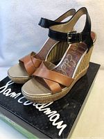 Side angle of Sam Edelman women's wedge sandal on box showing tan leather criss-cross straps, black ankle strap with buckle, and cork wedge heel.