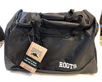 Front view of black Roots duffle bag with logo and attached tags