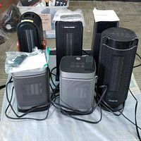 Photo showing six PTC ceramic heater fan ventilator mini tower units on cloth. Accessories such as remote controls and manuals visible. Various brands including Oraimo and PARIS RHÔNE visible on the front of units.