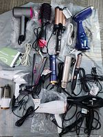 Overview of 19 hair styling tools laid out on fabric, including hair dryers, curling irons, and electric brushes with power cords visible.
