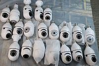 Top view of 20 white bulb shaped security cameras, some with dual axis motorized heads, placed in 4 rows on gray fabric backdrop.