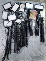 Overview of 9 LED light panels arranged with their tripods, cords, a sealed pack of color filter gels, and a wired remote controller visible on a grey surface.