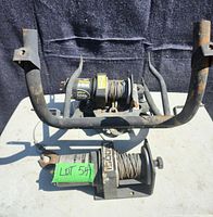 Front view of two 1500lb 12 volt winches, one mounted on a rusted metal ATV bumper, showing cables and hooks wound on spools.