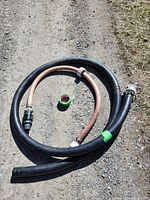 Full view of coiled black 2-inch hose with attached fittings plus smaller peach semi-rigid hose and short black hose segment, laid on gravel and dirt ground.