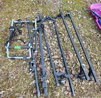 Photo showing trunk-mounted bike rack with straps alongside three separate sets of black metal roof rack crossbars. Placed on outdoor ground with leaves and grass.