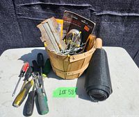Wide shot of hardware lot including wooden basket with metal brackets, pegboard hooks in packaging, screwdrivers lying on table, and rolled screen with wooden handle.