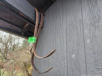 Two large rusted metal hooks hanging from a dark wooden beam outside, showing weathered grey siding in background.