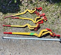 Four yellow load chain binders with red hooks laid on ground next to measuring tape showing lengths around 69-73 inches, visible wear and under sunlight.