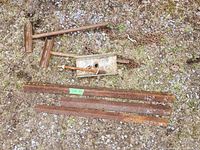Photo showing rusty L-shaped steel rods with handles, rectangular steel container with spout and handle, rusty chains, and three rusty rectangular metal bars of different lengths with size markings on gravel surface.