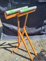 Pair of orange folding roller stands with silver metal rollers, standing on gravel surface in front of black backdrop. Photo taken to show overall condition and appearance.
