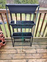 Full view of 3-tier black plastic planter boxes on a black metal frame with green metal trellis leaning in front.