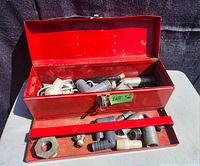 Open red metal toolbox showing various plumbing fittings including metal and plastic elbow joints, connector nuts, and pipe fittings inside and in front of the toolbox.