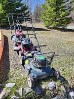 Three gas powered hand mowers shown outdoors on grass near trees.