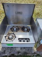 Front open view of stainless steel 3 burner propane stove showing burners, control knobs, and hinged lid with warning label.