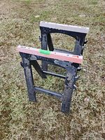 Overview of two black plastic sawhorses positioned outdoors on grass, showing significant paint splatter and wear.
