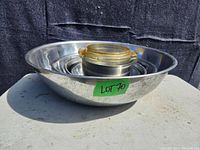 Six stainless steel nesting bowls including a small casserole dish with glass lid placed inside the largest bowl on a neutral outdoor surface.