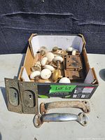 Full box containing various vintage door hardware pieces, including white ceramic knobs, brass lock inserts, and metal door handles.