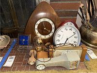 Photo of all clocks and barometer arranged on brick surface near metal fireplace screen. Shows Sessions wooden wall clock, blue desktop clock, tabletop barometer with glass dome, plastic wall clocks red and white, and other clocks.