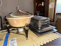 Side view of Fire King chaffing dish with lid on stand and silver plated casserole with trays stacked next to them on a lace tablecloth.