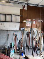Wide view showing assortment of garden tools including axes, shovels, pitch fork, snow stakes, tree trimmer and hand tools leaning against a wall and a ladder hanging above.