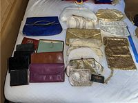 Overview of the assortment of handbags and wallets laid out on a white quilted surface, includes satin, leather, metal mesh and beaded styles.