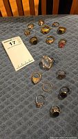 16 rings arranged on a blue textured cloth, including gold plated 18K marked rings, rings with various stones and one missing stone.
