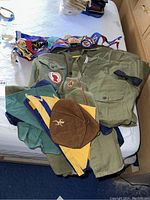 Photo showing an assortment of Boy and Cub Scout scarves, caps, shirts with patches, vintage pennants, and a black bow tie on a bed.