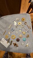 Top-down view of 27 assorted pins laid out on a gray fabric surface, showing variety of sizes, shapes, and designs with many rhinestone decorations.