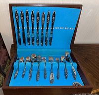 Full view of Oneida stainless flatware set inside blue-lined wooden case, showing knives, forks, and spoons arranged by type.