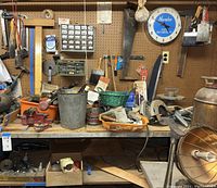 Workbench area with a variety of tools, including two red vices clamped to the table, hand tools hanging on pegboard, paint brushes, a gray metal bucket, and small parts organizers.