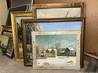 Four framed oil paintings leaning against pegboard, showing front winter village scene and three others behind