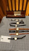 Full display of 8 assorted watches on a cloth surface, including a boxed Elgin watch and a variety of strap materials and watch face styles.