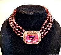Front view of the necklace displayed on a black bust, showing three strands of faceted purple crystal beads and the large rectangular purple crystal pendant framed by smaller crystals in antique gold tone metal.