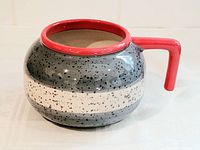 Front side view of large curling rock mug showing speckled gray and white bands with red handle and rim.