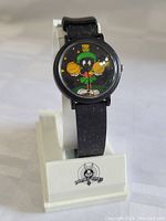 Front view of Marvin The Martian watch on stand showing character on black dial with stars, black strap, white display base with Looney Tunes logo