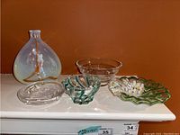 Photo showing five decorative art glass pieces arranged on a white surface: a heart-shaped piece, clear glass bowl, two floral-shaped bowls with colored accents, and a Sydenstricker bowl.