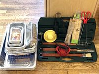 Set of foil food pans stacked on the left with grilling tools and accessories on the right inside an open green plastic carrying case, showing most items included in the lot.