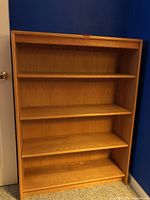 Front view of wood finish bookcase showing four shelves and light wood grain finish against blue wall.