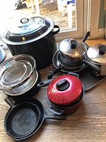 Assorted cookware including cast iron skillets, enamel pot, strainers, and mixing bowls on wooden floor.
