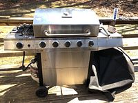 Full view of the stainless steel gas grill on wheels with propane tank and black cover beside it, showing overall condition and included items