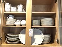 Full view of several shelves filled with Mikasa fine china including dinner plates, salad plates, cups, saucers, sugar bowl and creamer in white with floral designs.