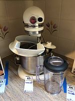 Front view of KitchenAid Model K5SS stand mixer with stainless steel bowl and clear plastic measuring beaker on counter.