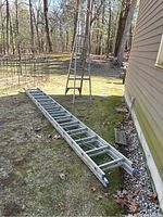 Photo showing both the 28ft metal extension ladder fully retracted on the ground and the 6ft metal step ladder standing upright outdoors on grass next to a house.