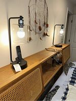 Pair of black metal table lamps with cylindrical clear glass shades on wooden surface beneath a wall hanging. Each lamp base has a wireless charger pad and a phone remote placed beside one.
