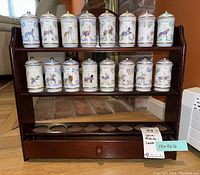 Full front view of wooden spice rack carousel with two shelves holding porcelain spice jars with carousel horse designs and gold knobs on lids. Empty base shelf with holes for jars and drawer knob visible.