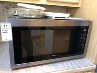Full view of Whirlpool microwave oven on counter showing stainless steel finish and black window door. Accessories including glass dish and paper plates on top.